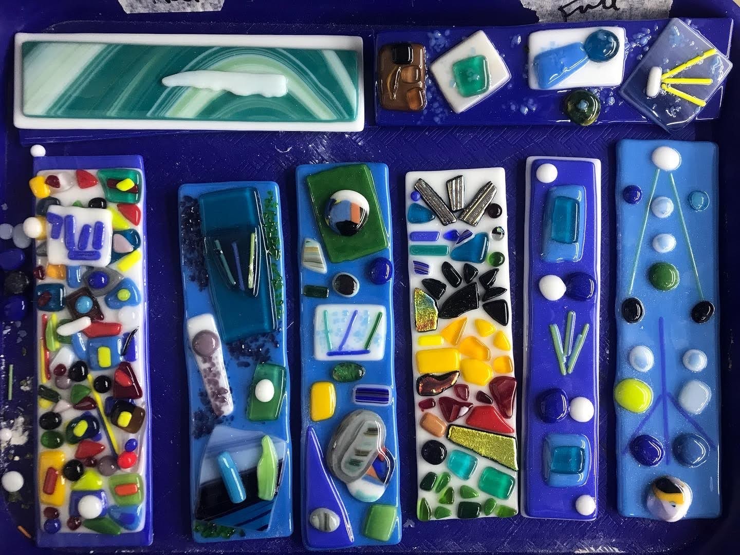 Colorful fused glass mezuzah covers and pieces laid out on a blue surface